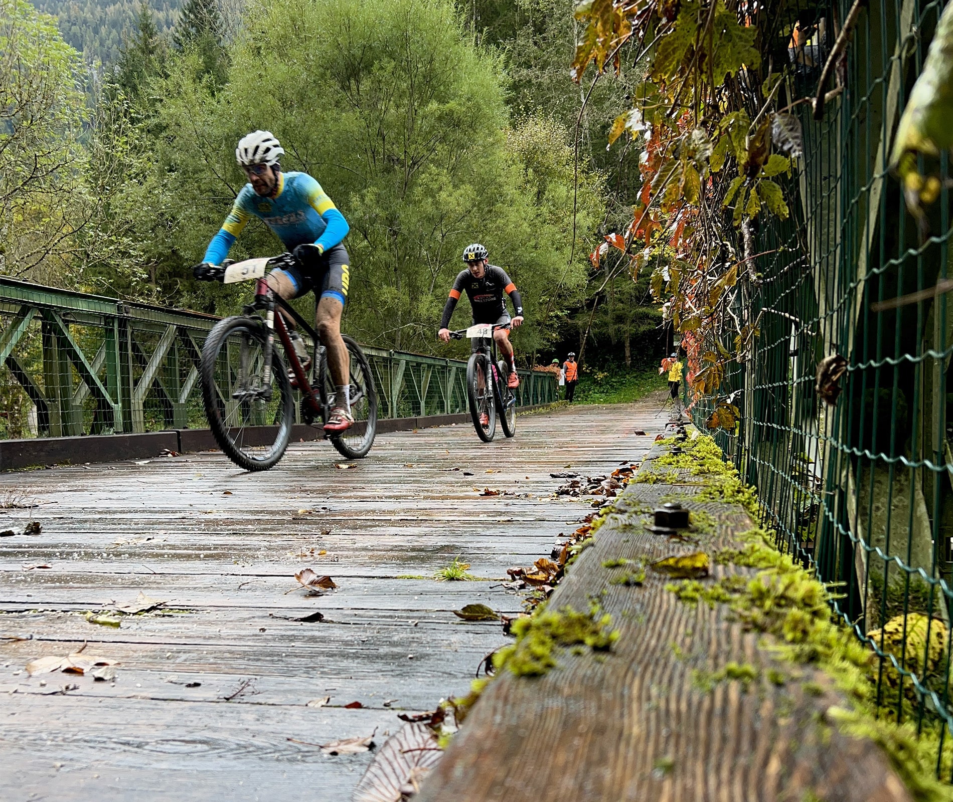 two mountainbikers chasing each other over a wooden bridge in wet autumn conditions, with race numbers attached to their bikes